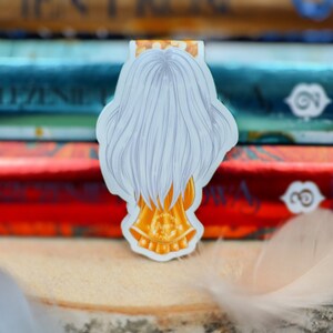 Puede incluir: Un marcap&aacute;ginas blanco y dorado con un personaje con el pelo largo blanco y una corona dorada. El marcap&aacute;ginas est&aacute; encima de una pila de libros.