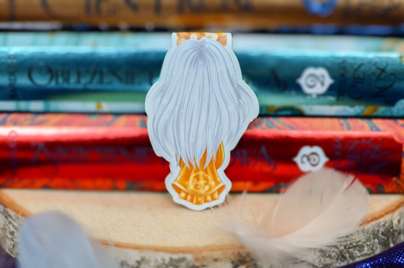 Puede incluir: Un marcap&aacute;ginas blanco y dorado con un personaje con el pelo largo blanco y una corona dorada. El marcap&aacute;ginas est&aacute; encima de una pila de libros.