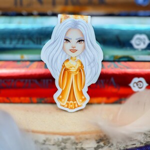 Puede incluir: Una mujer con cabello blanco, con una corona dorada y un vestido amarillo, sostiene un orbe dorado en sus manos. El fondo es una pila de libros.