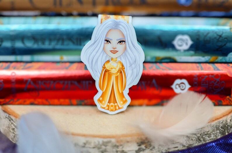 Puede incluir: Una mujer con cabello blanco, con una corona dorada y un vestido amarillo, sostiene un orbe dorado en sus manos. El fondo es una pila de libros.