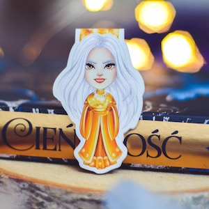 Puede incluir: Un marcap&aacute;ginas blanco y dorado con una ilustraci&oacute;n de dibujos animados de una mujer con el pelo largo y blanco que lleva una corona dorada y un vestido amarillo. El marcap&aacute;ginas est&aacute; encima de un libro con el t&iacute;tulo "Cień i Ośc" en letras doradas.