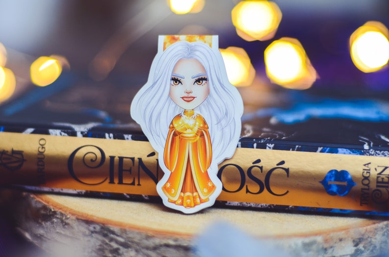 Puede incluir: Un marcap&aacute;ginas blanco y dorado con una ilustraci&oacute;n de dibujos animados de una mujer con el pelo largo y blanco que lleva una corona dorada y un vestido amarillo. El marcap&aacute;ginas est&aacute; encima de un libro con el t&iacute;tulo "Cień i Ośc" en letras doradas.