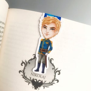 Puede incluir: Un marcapáginas con una ilustración de dibujos animados de un joven con cabello rubio que lleva un uniforme militar azul y dorado. El marcapáginas tiene el nombre "Nikolaj" impreso en la parte inferior.