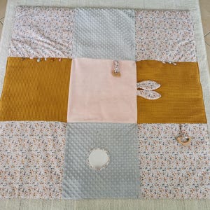 Könnte beinhalten: Eine Baby-Spielmatte mit Patchwork-Design. Die Matte hat Abschnitte aus senfgelben, hellrosa und grauen Stoffen sowie einen Blumendruck. Sie enthält Holzspielzeug und einen Spiegel.