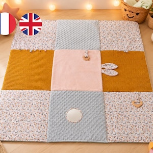 Peut inclure: Tapis d'éveil pour bébé aux couleurs vives. Il présente un motif patchwork avec des sections florales, jaune moutarde, rose clair et tissu gris texturé. Il comprend des jouets en bois et un petit miroir pour l'éveil sensoriel.
