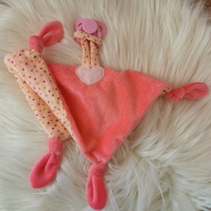 Puede incluir: Una manta de seguridad para bebé de color coral suave con un chupete adjunto. La manta presenta una aplicación en forma de corazón y dos brazos estampados con lunares rosas. El chupete y los bordes de la manta son de color coral a juego.