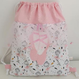 Puede incluir: Mochila rosa con cordón y estampado de bailarina. La bolsa presenta una gran zapatilla de ballet bordada y un cierre de cordón rosa. El diseño incluye ilustraciones de bailarinas, estrellas y formas geométricas. Ideal para material de ballet.