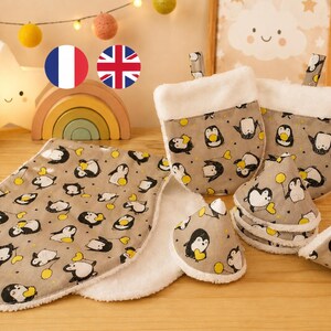 Puede incluir: Un juego de regalo para bebé con babero, manoplas, gasas y mordedor. Los artículos están estampados con dibujos de pingüinos en negro, blanco y amarillo sobre un fondo beige. El juego incluye un ribete blanco y esponjoso.
