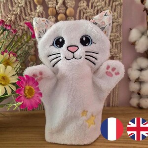 May include: A white cat-shaped hand puppet with embroidered facial features, including blue eyes, a pink nose, and black whiskers. The puppet has pink paw prints and yellow star accents. The ears have a floral pattern.