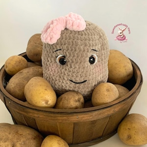 May include: A crocheted potato character with a pink bow, smiling eyes, and a basket of potatoes. The plush potato is light brown and surrounded by several real potatoes in a wooden basket. The image has a soft, inviting aesthetic.