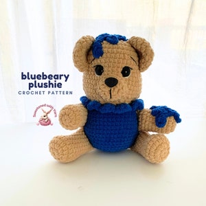 May include: Crochet pattern for a blue and brown teddy bear plush toy. The bear is wearing a blue shirt with a ruffled collar. The pattern is called "bluebeary plushie".