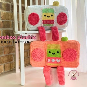 May include: Two crocheted boombox plushies, one white with pink and green details, and one orange with pink and green details. Both have smiling faces and are sitting on a white chair. The text "boombox plushie crochet pattern" is visible at the bottom of the image.
