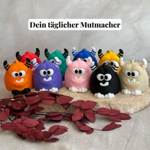 Könnte beinhalten: Eine Sammlung farbenfroher Monster-Schlüsselanhänger. Jeder Schlüsselanhänger hat eine einzigartige Farbe, darunter Orange, Lila, Rosa, Schwarz und Gelb. Sie haben weiße Augen, einen lächelnden Mund und kleine Hörner mit schwarz-weißen Streifen. Der Text "Dein täglicher Mutmacher" ist oben zu sehen.