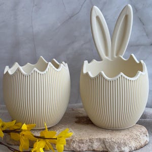 Puede incluir: Dos macetas de cerámica color crema con forma de huevo de Pascua. Una maceta tiene orejas de conejo. Ambas tienen un borde festoneado y líneas verticales. Flores amarillas en primer plano. Las macetas están sobre una superficie de piedra.