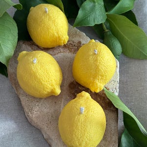 May include: Four yellow lemon-shaped candles with white wicks are arranged on a light brown stone. Green leaves and a small green lemon are in the background. The candles have a textured surface.
