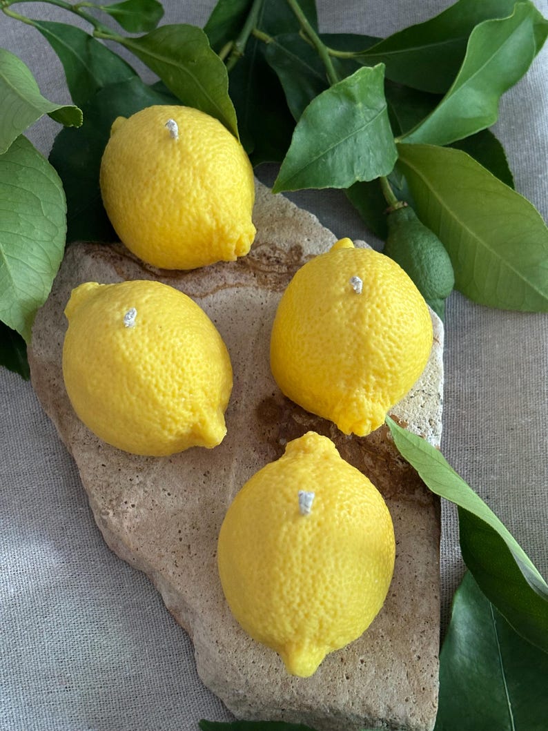 May include: Four yellow lemon-shaped candles with white wicks are arranged on a light brown stone. Green leaves and a small green lemon are in the background. The candles have a textured surface.