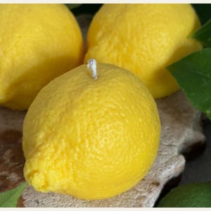 May include: Three bright yellow lemon-shaped candles. One candle is in focus, with a white wick. The candles have a textured surface, mimicking the appearance of real lemons. Green leaves are visible in the background.
