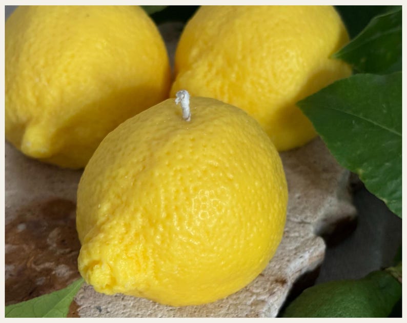 May include: Three bright yellow lemon-shaped candles. One candle is in focus, with a white wick. The candles have a textured surface, mimicking the appearance of real lemons. Green leaves are visible in the background.