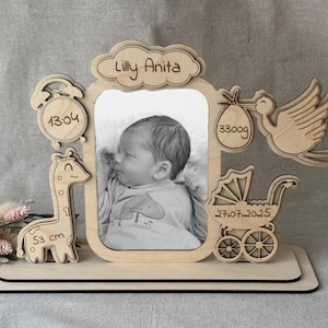 Può includere: Cornice portafoto per bambini in legno con foto di un neonato. La cornice include una nuvola con il nome "Lilly Anita", un orologio, una giraffa, una cicogna che trasporta un fagotto e un passeggino. La giraffa è alta 53 cm e il bambino pesava 3300g.