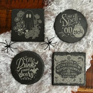 Op de afbeelding: Vier zwarte leistenen onderzetters met Halloween-ontwerpen. Eén vierkante onderzetter toont een spook omringd door bloemen. De andere onderzetters zijn rond en tonen de zinnen "I Put a Spell on You" en "Eat, Drink and Be Scary". De vierde onderzetter is een Ouija bord.