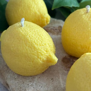 May include: Close-up of four yellow lemon-shaped candles with white wicks. The candles have a textured surface, mimicking the appearance of real lemons. The candles are arranged on a light brown, textured surface, with green leaves in the background.