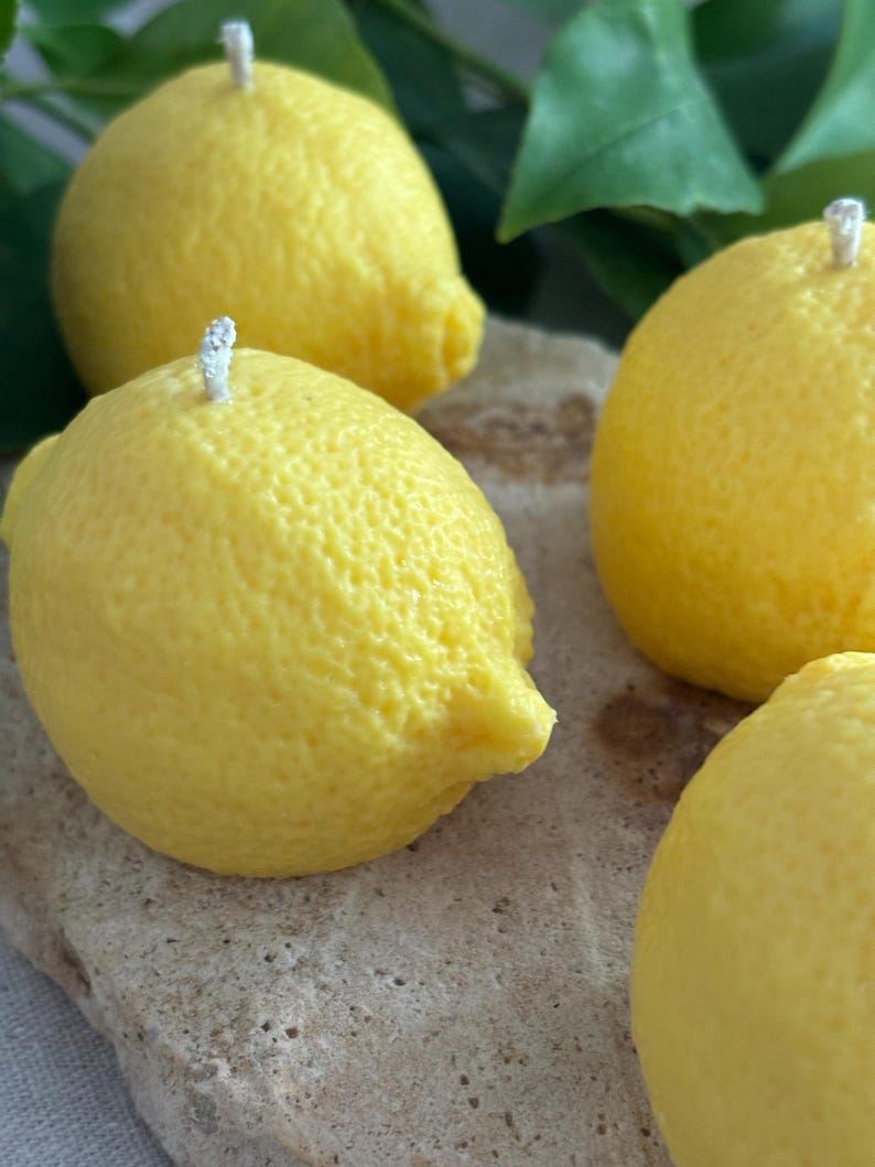 May include: Close-up of four yellow lemon-shaped candles with white wicks. The candles have a textured surface, mimicking the appearance of real lemons. The candles are arranged on a light brown, textured surface, with green leaves in the background.