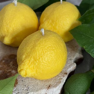 May include: Three yellow lemon-shaped candles with white wicks are arranged on a textured, light-colored surface. Green leaves and small green lemons surround the candles, creating a fresh, natural aesthetic. The candles have a realistic, textured appearance.