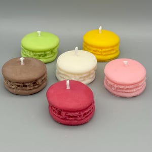 Peut inclure: Six bougies rondes en forme de macarons, de couleurs variées : vert, jaune, blanc, rose, marron et rouge. Chaque bougie a une mèche et un design texturé et superposé sur les côtés. Elles sont disposées sur une surface grise.