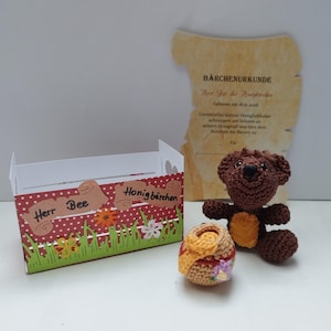 Könnte beinhalten: Ein handgehäkelter Teddybär mit einem kleinen Honigtopf und einer dekorativen Box. Die Box ist rot mit weißen Punkten, grünem Gras und den Worten "Herr Bee" und "Honigbärchen". Eine urkundenförmige Schriftrolle befindet sich im Hintergrund.