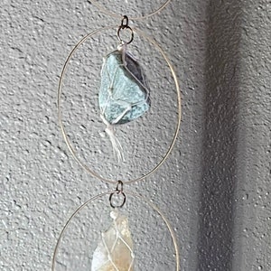 May include: A hanging mobile featuring various colorful crystals, each encased in a white net and suspended within gold-colored metal rings. The crystals include pink, blue, green, yellow, and red stones.
