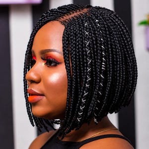 May include: A black bob-length braided wig featuring a centre part and small, uniform box braids. The hairstyle includes silver metallic hair cuffs and decorative silver thread accents woven into several of the braids, creating a textured look.