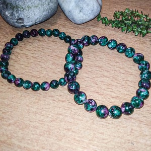 May include: Two green and purple gemstone bracelets with round beads. The bracelets are on a wooden surface.