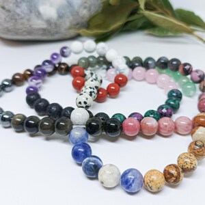 Pode incluir: Uma cole&ccedil;&atilde;o de pulseiras de contas em v&aacute;rias cores, incluindo preto, roxo, vermelho, rosa e azul. As contas redondas parecem ser pedras preciosas. As pulseiras est&atilde;o dispostas em um padr&atilde;o semicircular.