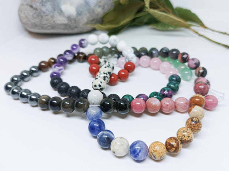 Pode incluir: Uma cole&ccedil;&atilde;o de pulseiras de contas em v&aacute;rias cores, incluindo preto, roxo, vermelho, rosa e azul. As contas redondas parecem ser pedras preciosas. As pulseiras est&atilde;o dispostas em um padr&atilde;o semicircular.