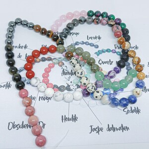 Pode incluir: Uma cole&ccedil;&atilde;o de pulseiras de contas em v&aacute;rias cores e tipos de pedra, incluindo hematita, quartzo e sodalita. As contas s&atilde;o enfiadas, mostrando uma variedade de texturas e tons, de pretos profundos a azuis e rosas vibrantes.