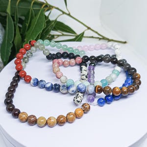 May include: A selection of beaded bracelets in diverse colours, such as red, green, pink, black, blue, and brown. The bracelets are crafted from various stones and presented on a white surface. Green leaves are visible in the background.