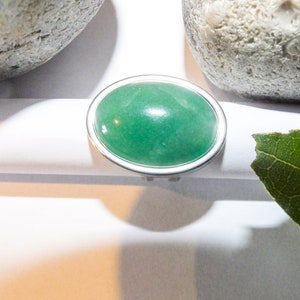 Puede incluir: Un anillo de plata con una gema verde ovalada grande. El anillo está sobre un fondo blanco, con dos piedras grises y una hoja verde en el fondo. La gema tiene una superficie lisa y pulida.