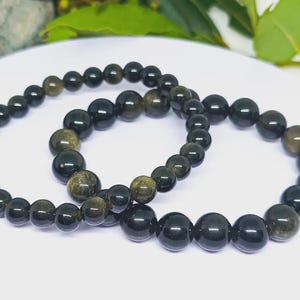 Peut inclure: Deux bracelets de perles avec des perles noires et dorées. Les bracelets sont composés de perles rondes polies, présentés sur une surface blanche. Les perles ont une finition brillante et l'or ajoute un léger scintillement. Les perles mesurent environ 0,8 cm de diamètre.