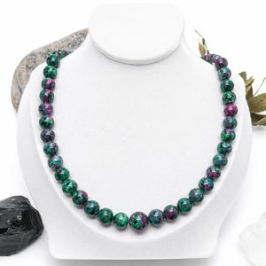 Peut inclure: Un collier de perles composé de pierres rondes et polies présentant un motif marbré de vert forêt profond et de rose magenta vif. Les perles sont de taille uniforme, enfilées pour former un cordon circulaire continu sur un présentoir blanc.