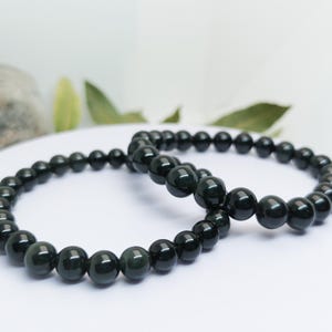 Può includere: Due braccialetti elastici composti da perline sferiche in ossidiana nera, lisce e levigate. Le perline presentano una superficie lucida e riflettente con sottili sfumature verde scuro visibili sotto la luce, disposte in cerchio.
