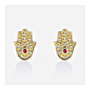 Peut inclure: Une paire de boucles d'oreilles en or en forme de main de Hamsa, un symbole de protection. Chaque boucle d'oreille est ornée de petites pierres de zircone cubique blanches et d'un seul rubis rouge au centre.