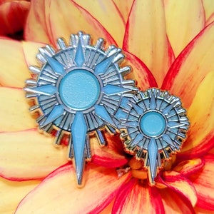 Könnte beinhalten: Zwei silberfarbene Emaille-Anstecknadeln mit einem hellblauen Sonnenstrahl-Design. Die Anstecknadeln haben einen zentralen Kreis und strahlenförmige Punkte, vor einem gelb-roten Blumenhintergrund. Die Anstecknadeln sind wahrscheinlich dekoratives Zubehör.