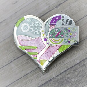 May include: A heart-shaped metal pin with a pink, green, and purple design. The pin features a gear, a speedometer, and hearts.