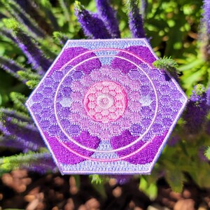 Puede incluir: Un parche hexagonal bordado de color morado y rosa con un diseño geométrico. El parche tiene un contorno blanco y un centro blanco.
