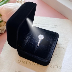 May include: A diamond engagement ring in a black velvet box. The ring features a large, round-cut diamond center stone and a band adorned with smaller diamonds. The box is open, revealing the ring against a dark interior.