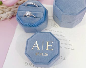 Puede incluir: Dos cajas octogonales de terciopelo azul claro para anillos. Una caja está abierta y contiene una alianza de diamantes y un anillo solitario. La otra caja está cerrada, con el texto dorado A | E y la fecha 07.11.26 en la tapa superior.