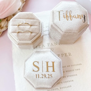 May include: Two octagonal beige velvet ring boxes with gold lettering. One box is open, displaying a gold solitaire ring and a gold chevron band. The other box is closed, featuring the name Tiffany. A third box shows the initials S|H and 11.29.25.