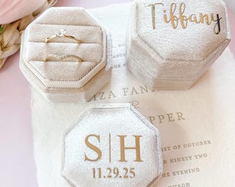 May include: Three beige velvet ring boxes, one open with two gold rings. Two boxes have gold lettering: "Tiffany" and "SH" with the date "11.29.25". The boxes are octagonal and sit on a cream-colored surface.