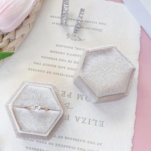 May include: A gold engagement ring with a round diamond is displayed in a gray velvet hexagonal ring box. Two diamond hoop earrings and another empty ring box are also visible on a vintage paper with text.