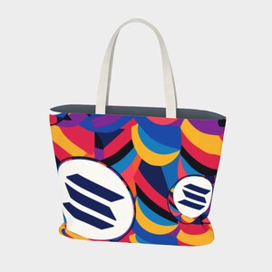 May include: A colorful tote bag with a geometric pattern in shades of red, yellow, blue, and purple. The bag has two white handles and features a white circle with a blue logo in the center.